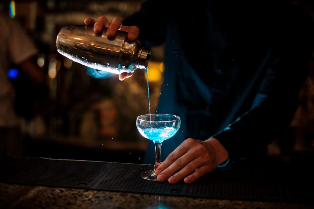 barman pours from a shaker into a glass of alcohol cocktail Blue Lagoon barman in a blue shirt pours from a shaker into a glass of alcohol cocktail Blue Lagoon on a dark background in the bar